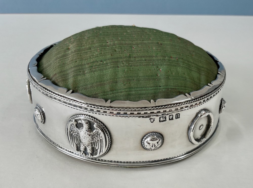 A circular Arts and Crafts silver pin cushion stand with embossed eagle medallions and a raised green woven fabric top. View II