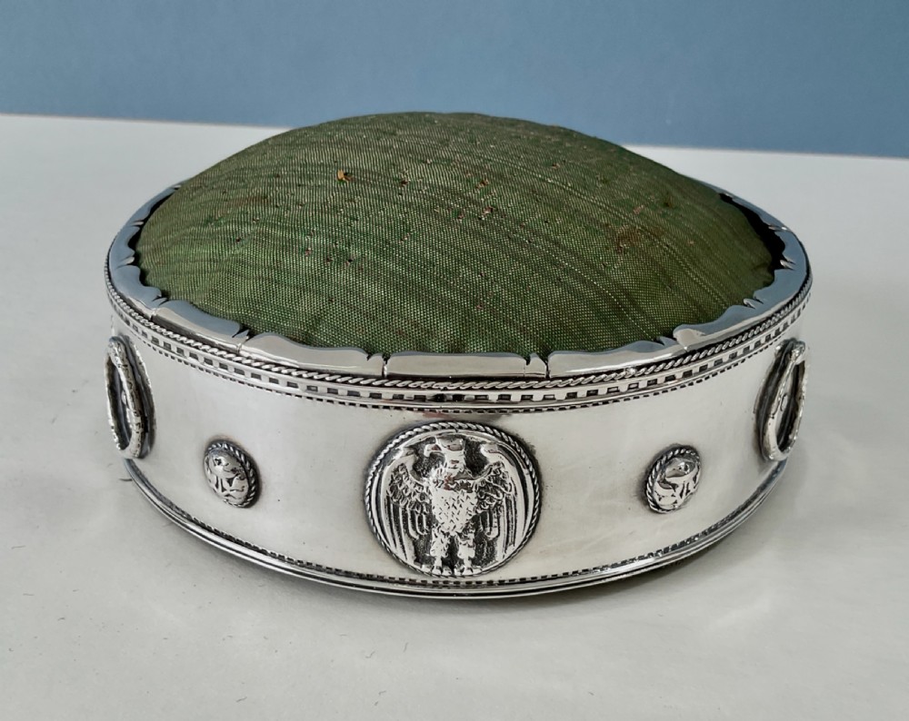 A circular Arts and Crafts silver pin cushion stand with embossed eagle medallions and a raised green woven fabric top. View V