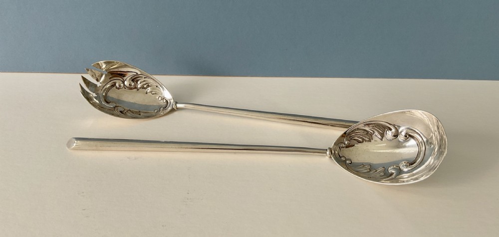 A pair of antique silver salad servers with long slender handles and embossed scrollwork on the spoon and fork bowls.