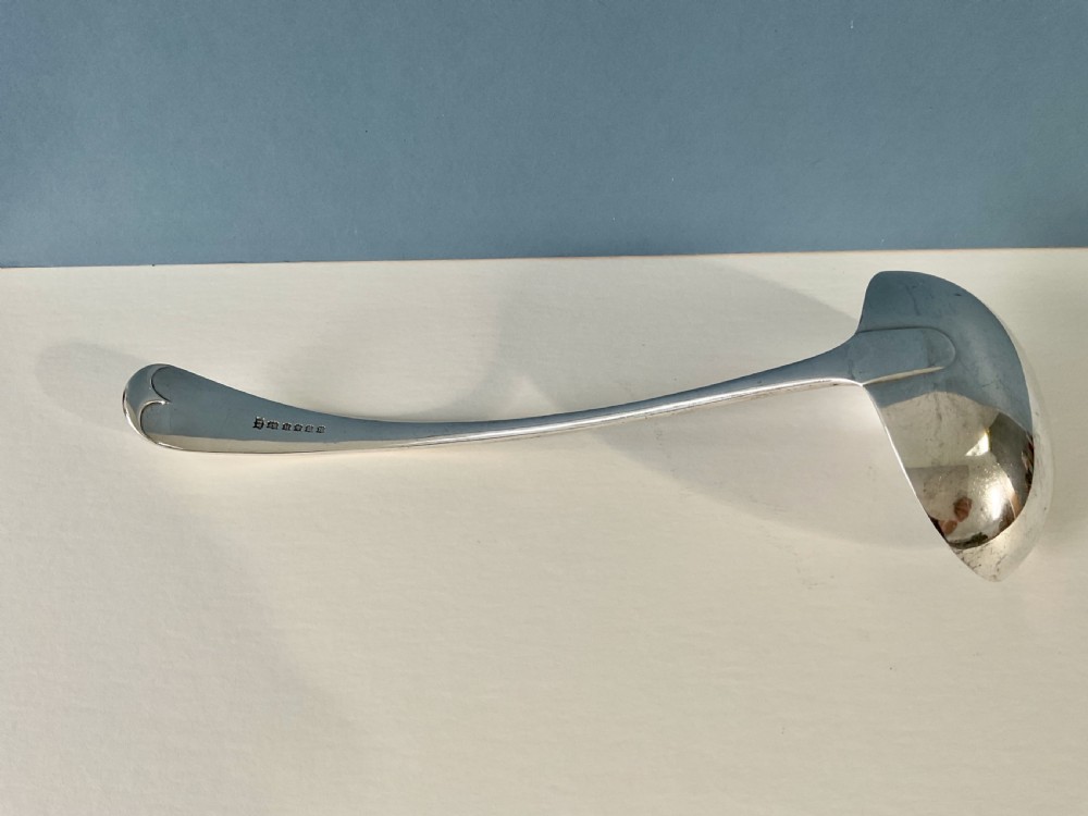 A silver-plated soup ladle featuring a deep hemispherical bowl and a long curved handle resting horizontally on a light surface. View V