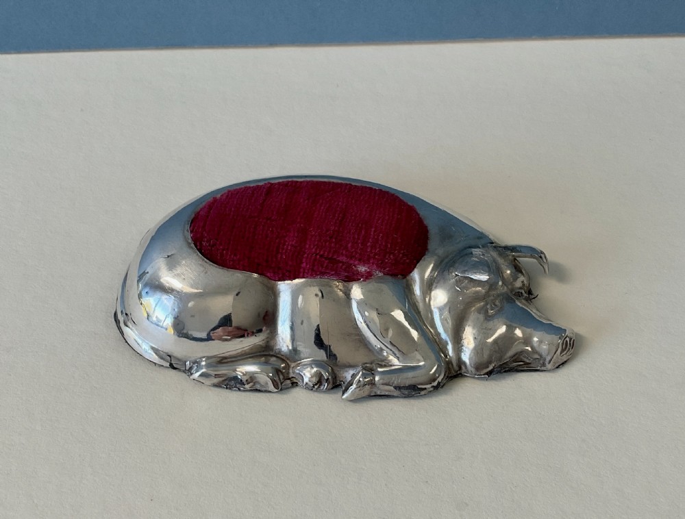 An antique silver pin cushion cast in the form of a recumbent pig with a red velvet cushion insert in its back.