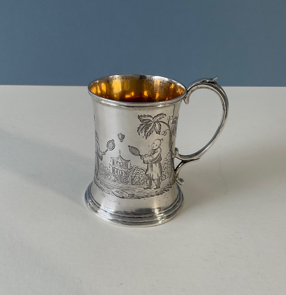 A silver mug featuring detailed Chinoiserie engravings of figures playing battledore and shuttlecock alongside a pagoda, fitted with a loop handle and a gold interior.