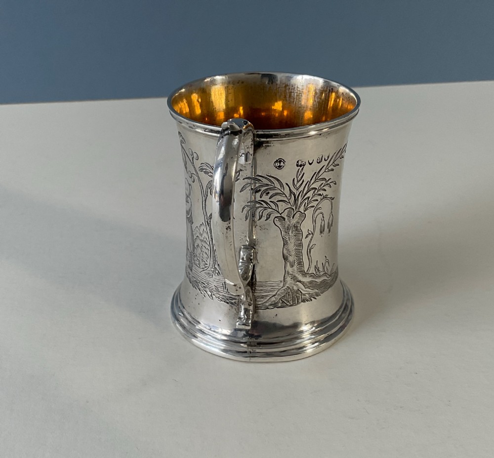 A silver mug featuring detailed Chinoiserie engravings of figures playing battledore and shuttlecock alongside a pagoda, fitted with a loop handle and a gold interior. View III