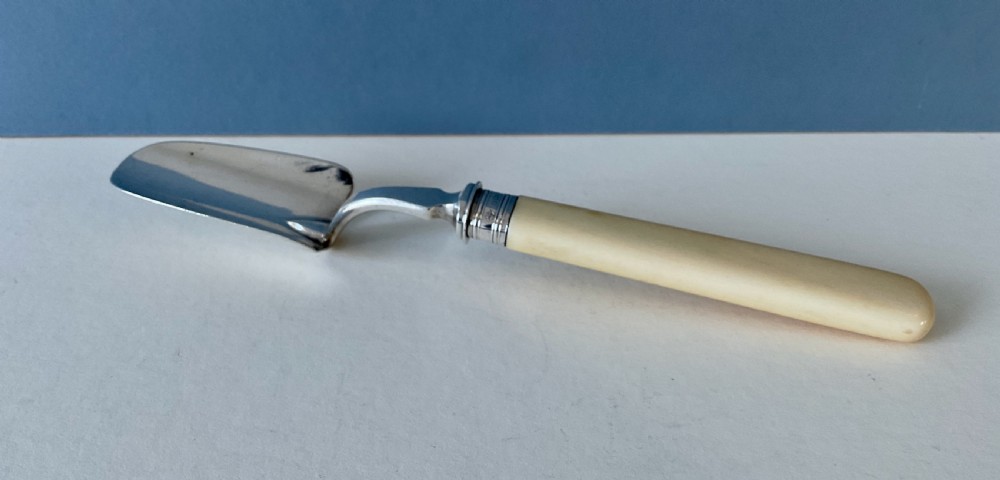 An antique silver cheese scoop featuring a curved spade blade, silver ferrule, and a smooth cream-coloured handle.