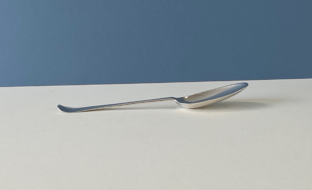 A single George I sterling silver spoon with a central ridge handle, upturned terminal, and an oval bowl against a split blue and white background. View V