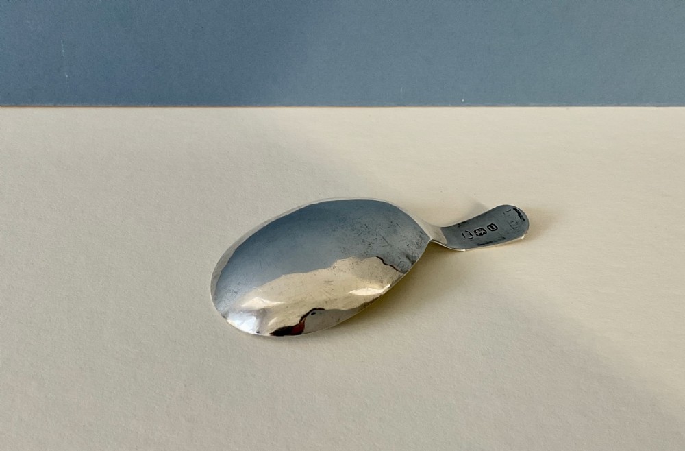 An antique Georgian sterling silver tea caddy spoon with a wide shallow bowl and a curved handle displayed on a light surface. View II