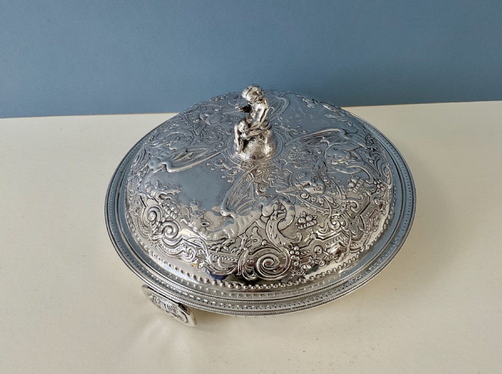 An ornate round Georgian solid silver entree dish featuring a domed lid decorated with repousse scrolling motifs, a figural finial, and a beaded rim.