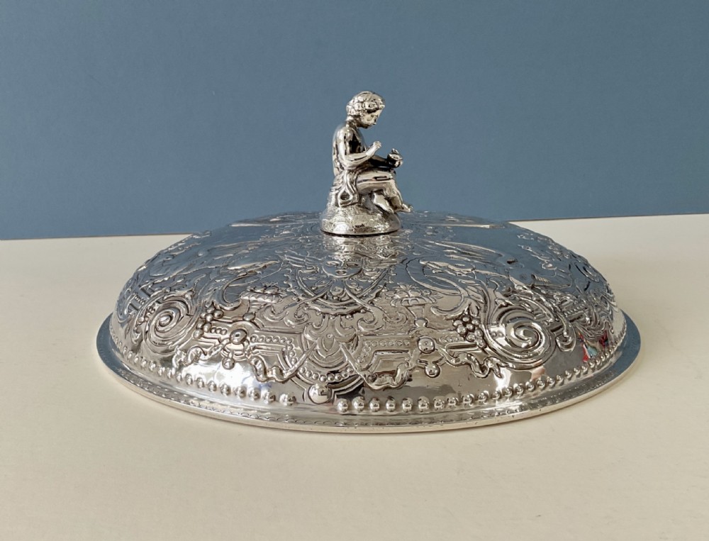 An ornate round Georgian solid silver entree dish featuring a domed lid decorated with repousse scrolling motifs, a figural finial, and a beaded rim. View II