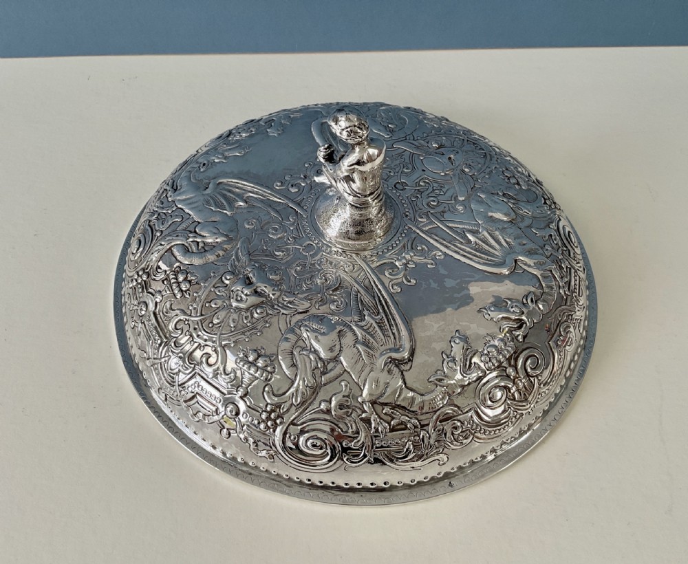 An ornate round Georgian solid silver entree dish featuring a domed lid decorated with repousse scrolling motifs, a figural finial, and a beaded rim. View III