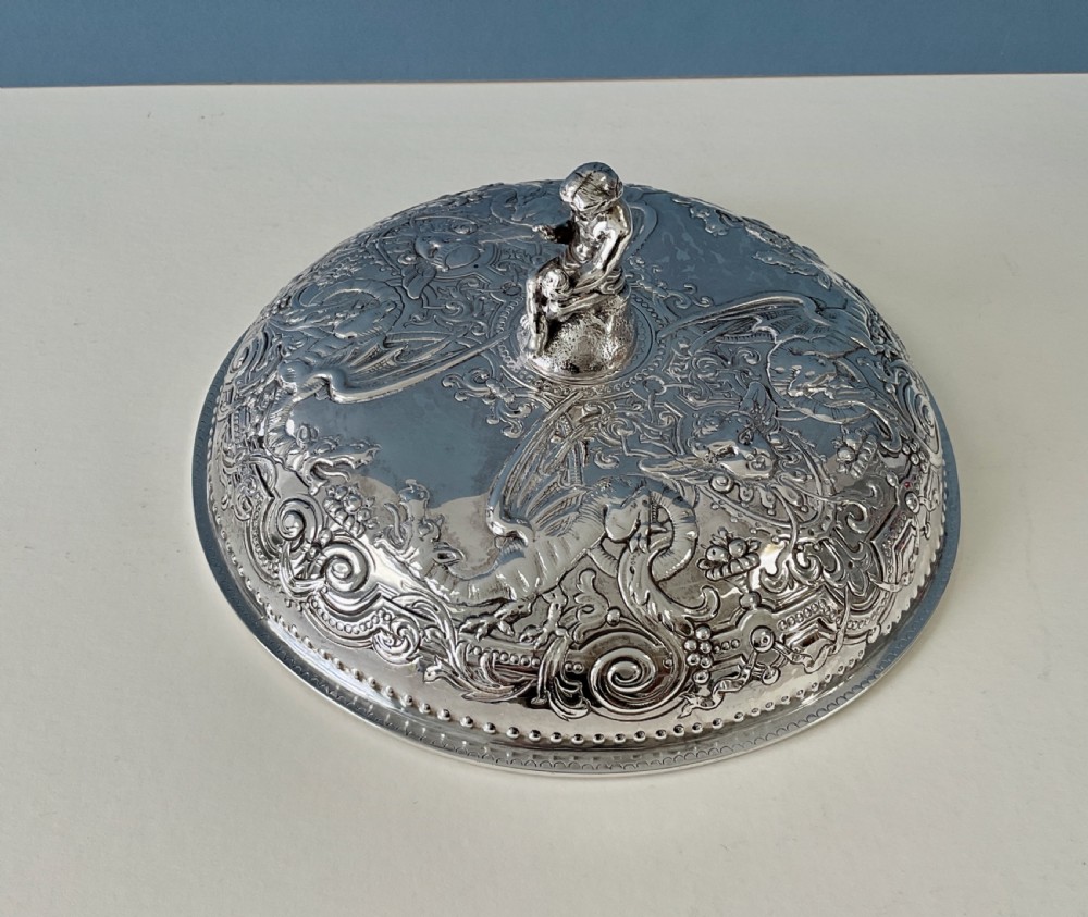 An ornate round Georgian solid silver entree dish featuring a domed lid decorated with repousse scrolling motifs, a figural finial, and a beaded rim. View V
