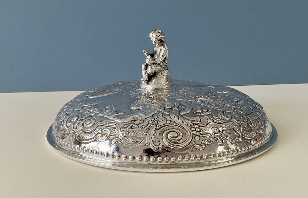 An ornate round Georgian solid silver entree dish featuring a domed lid decorated with repousse scrolling motifs, a figural finial, and a beaded rim. View VI