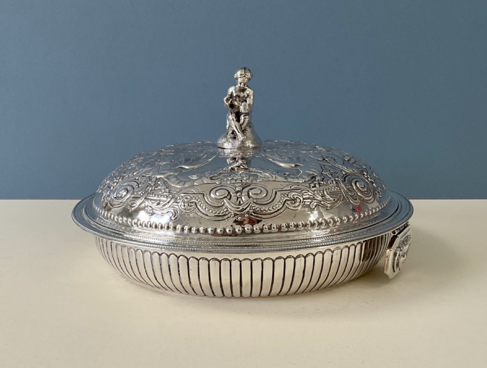 An ornate round Georgian solid silver entree dish featuring a domed lid decorated with repousse scrolling motifs, a figural finial, and a beaded rim. View X