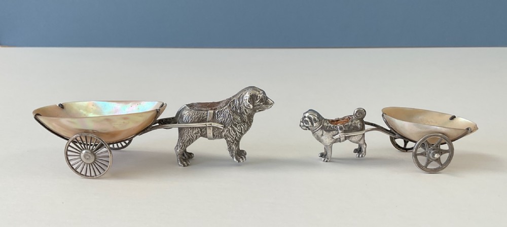 A pair of antique silver novelty pin cushions featuring detailed dog figures pulling mother-of-pearl shell carts on two wheels.