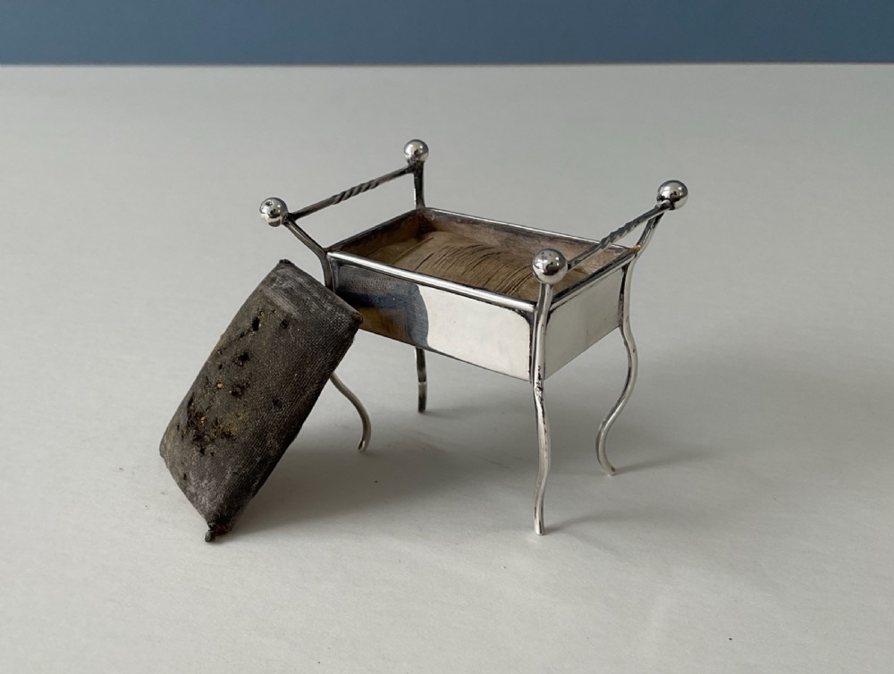 An antique silver pin cushion shaped like a miniature piano stool with slender curved legs and a dark velvet padded cushion. View IV