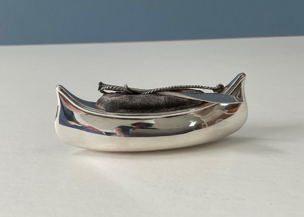 An antique silver canoe-shaped pin cushion featuring a central dark fabric cushion and miniature oar details.