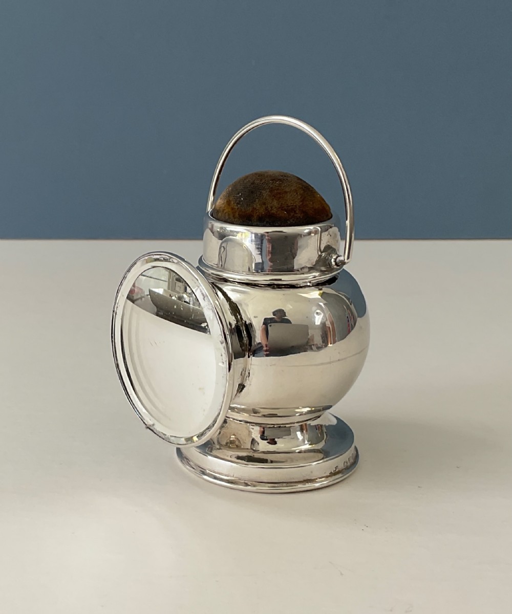 An antique silver novelty pin cushion shaped like a miniature railway lantern with a round convex lens and a dark fabric cushion top.