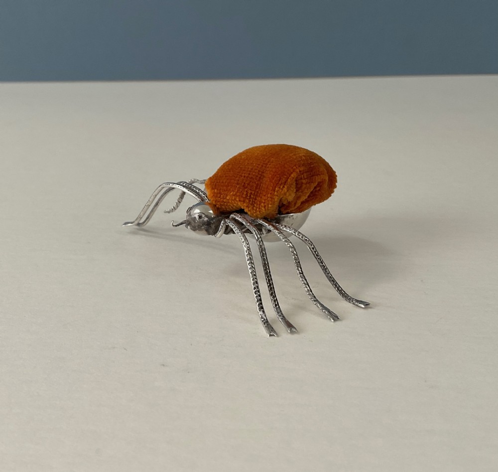 A silver pincushion cast in the form of a spider, featuring a dark orange velvet cushion on its back.