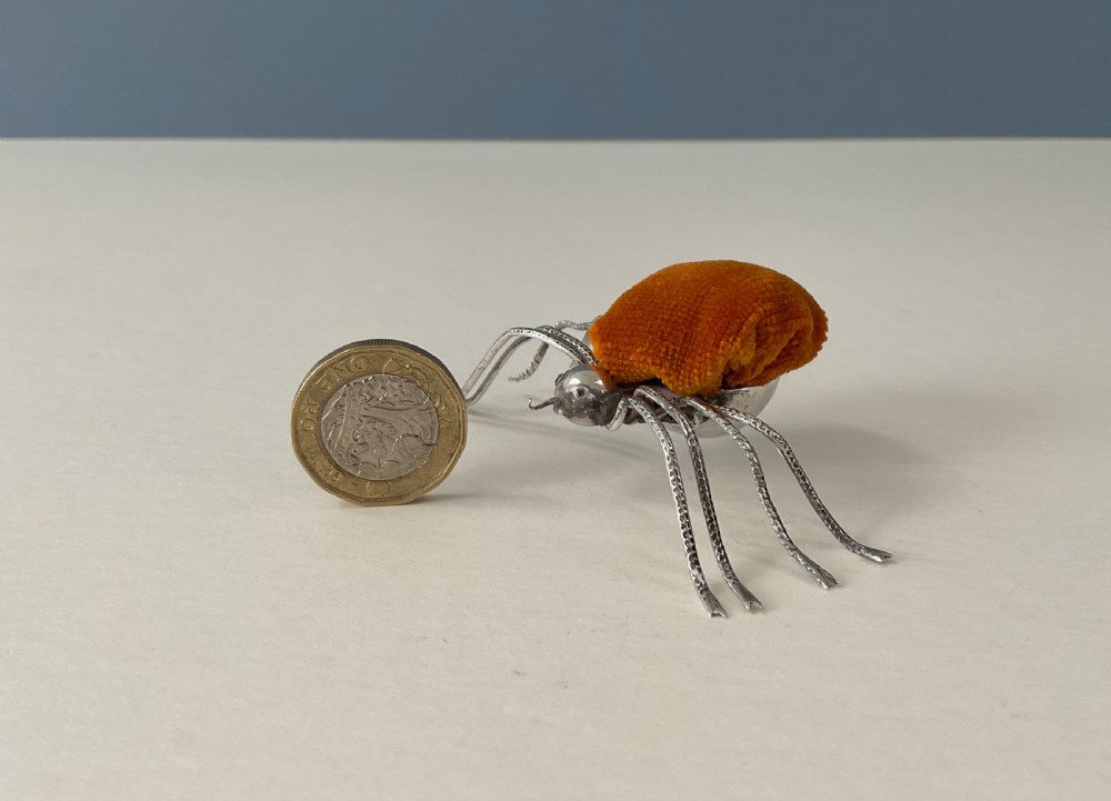 A silver pincushion cast in the form of a spider, featuring a dark orange velvet cushion on its back. View II