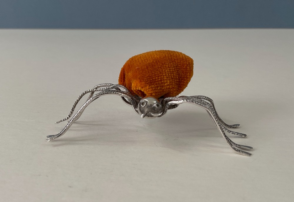 A silver pincushion cast in the form of a spider, featuring a dark orange velvet cushion on its back. View IV