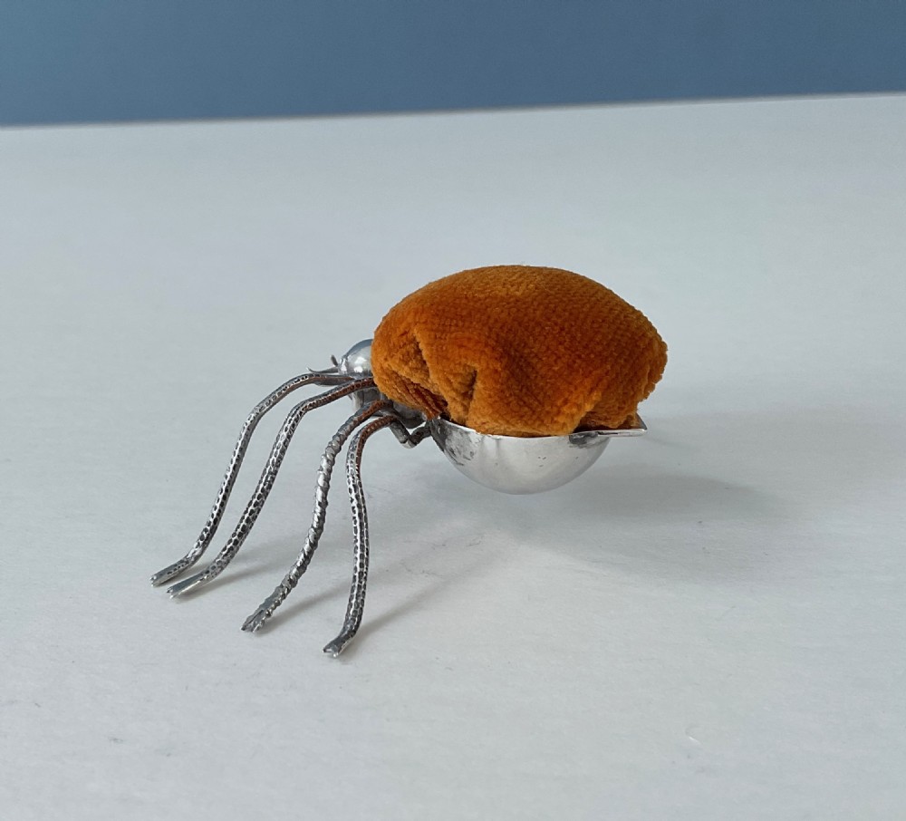 A silver pincushion cast in the form of a spider, featuring a dark orange velvet cushion on its back. View V