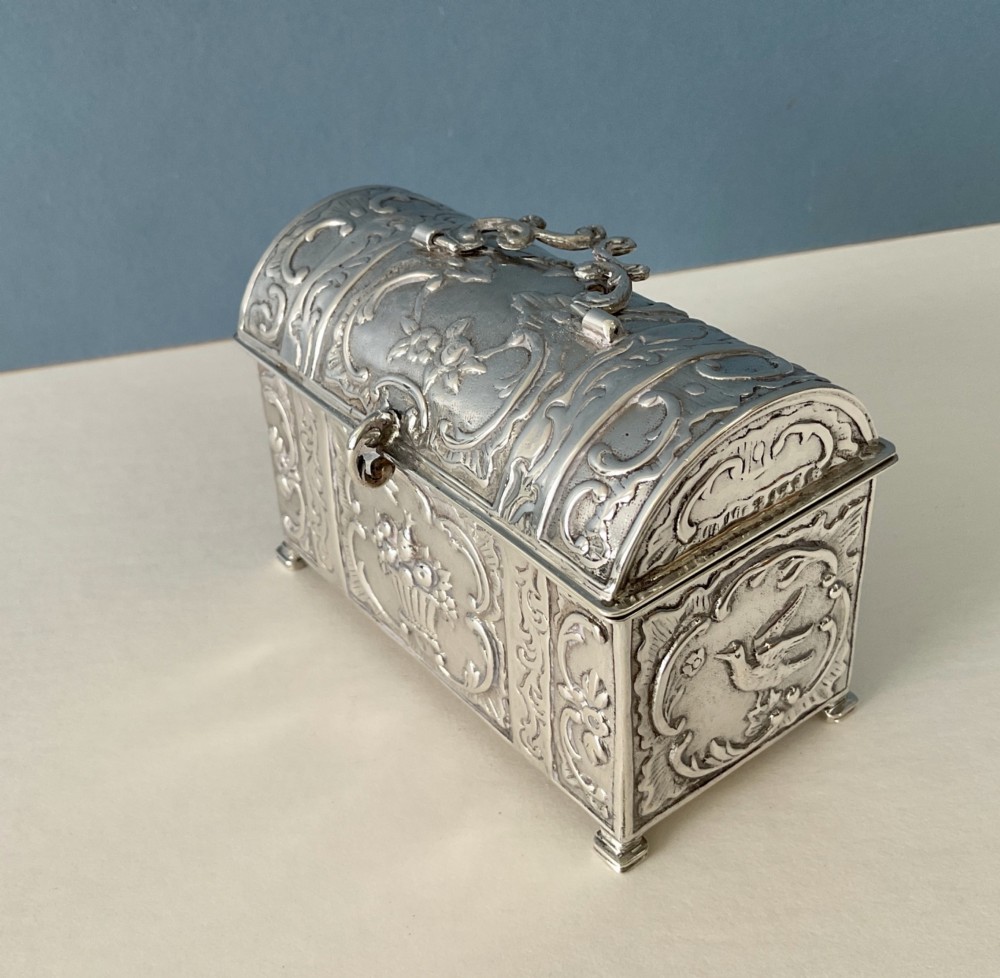 A small rectangular silver marriage casket featuring a domed lid, top handle, bracket feet, and intricate repousse scrollwork depicting birds and floral motifs.