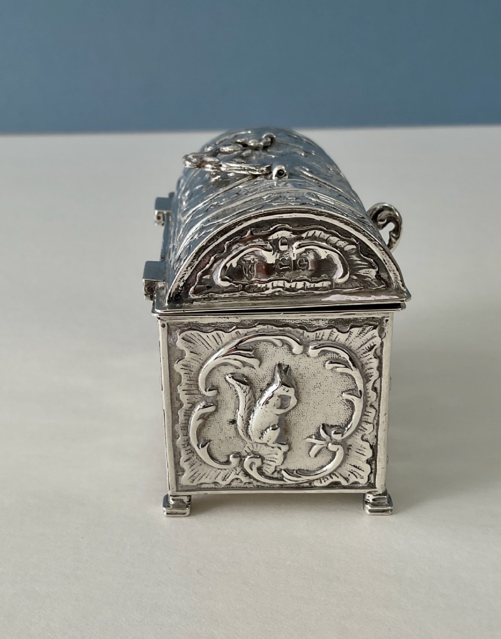 A small rectangular silver marriage casket featuring a domed lid, top handle, bracket feet, and intricate repousse scrollwork depicting birds and floral motifs. View V