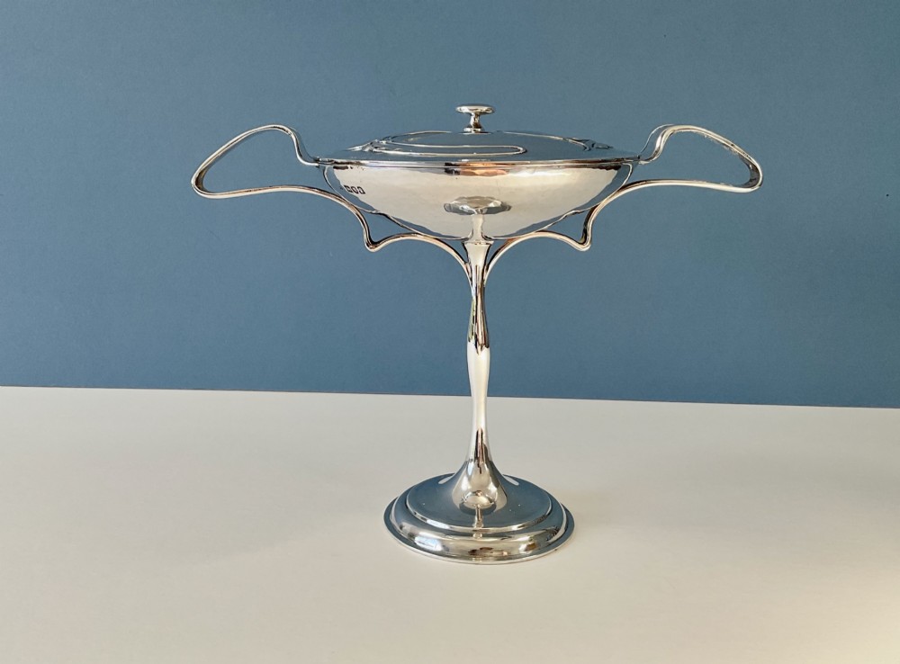 An Art Nouveau silver lidded bowl featuring high looping handles and an elongated pedestal stem against a plain blue background.
