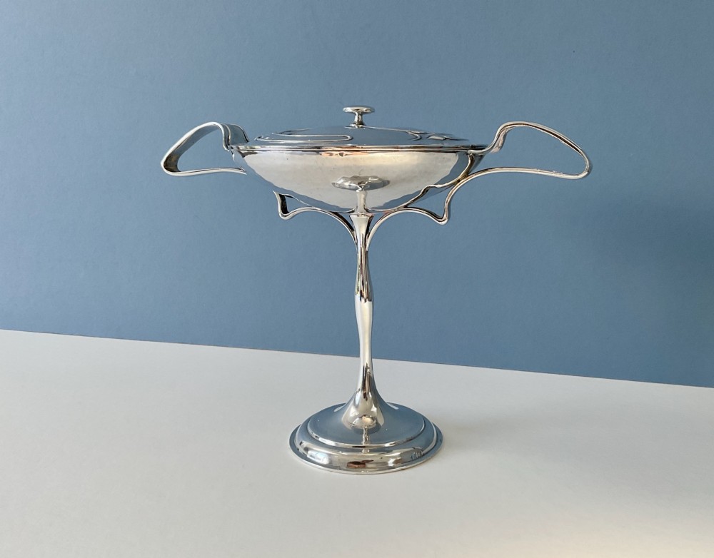 An Art Nouveau silver lidded bowl featuring high looping handles and an elongated pedestal stem against a plain blue background. View II