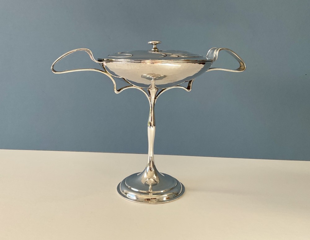 An Art Nouveau silver lidded bowl featuring high looping handles and an elongated pedestal stem against a plain blue background. View III