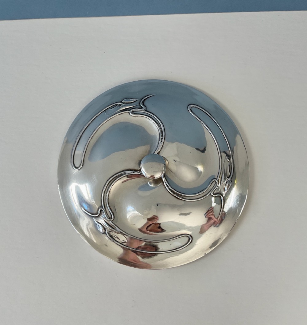 An Art Nouveau silver lidded bowl featuring high looping handles and an elongated pedestal stem against a plain blue background. View IV