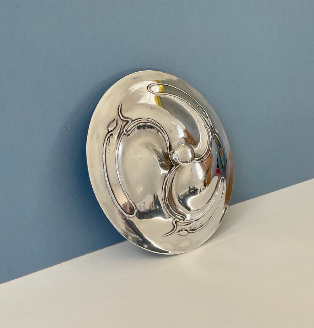 An Art Nouveau silver lidded bowl featuring high looping handles and an elongated pedestal stem against a plain blue background. View V