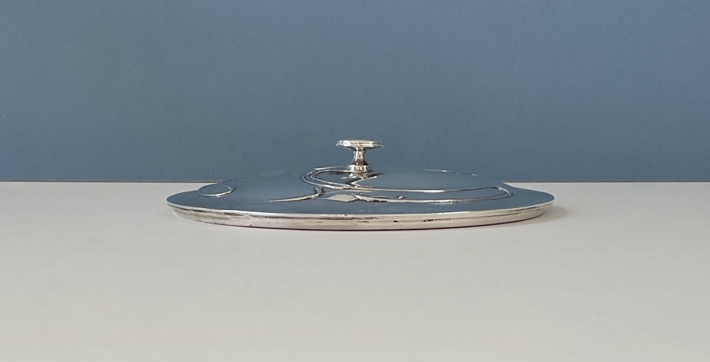 An Art Nouveau silver lidded bowl featuring high looping handles and an elongated pedestal stem against a plain blue background. View VII