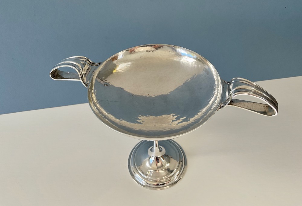 An Art Nouveau silver lidded bowl featuring high looping handles and an elongated pedestal stem against a plain blue background. View VIII
