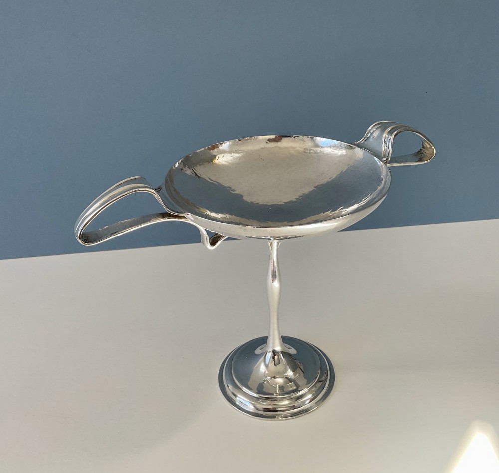 An Art Nouveau silver lidded bowl featuring high looping handles and an elongated pedestal stem against a plain blue background. View IX