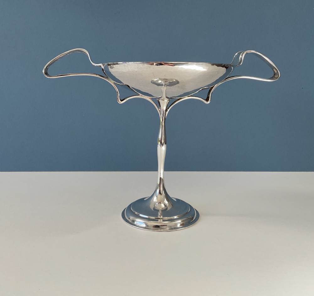 An Art Nouveau silver lidded bowl featuring high looping handles and an elongated pedestal stem against a plain blue background. View XI