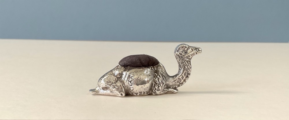 An antique silver novelty pin cushion cast in the form of a seated camel with a dark fabric cushion integrated into its back.