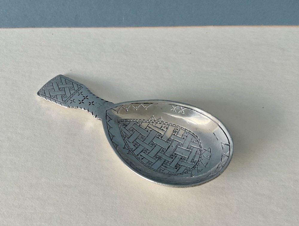 An antique sterling silver caddy spoon featuring a teardrop bowl and flat handle decorated with geometric basketweave engraving. View V