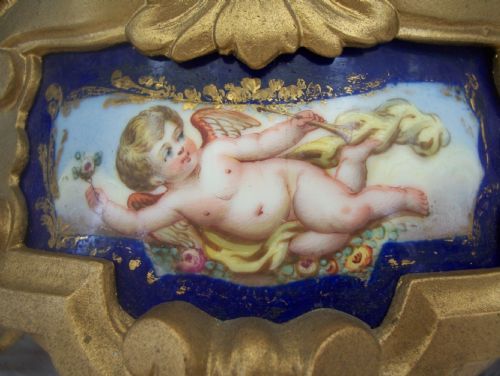 fig 1. An ornate 19th-century French gilt metal mantel clock featuring three sculpted putti figures surrounding a circular enamel dial and a painted porcelain plaque base.