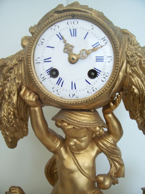 fig 2. An ornate 19th-century French gilt metal mantel clock featuring three sculpted putti figures surrounding a circular enamel dial and a painted porcelain plaque base.