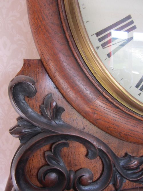 fig 1. An early nineteenth-century carved oak wall clock featuring a white circular dial with Roman numerals, protected by convex glass in a brass bezel, mounted on a carved wooden bracket base.
