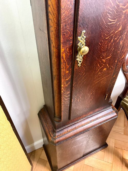 fig 1. An Early Georgian longcase clock constructed from dark oak featuring a square hood, brass dial with Roman numerals, and an arched trunk door.