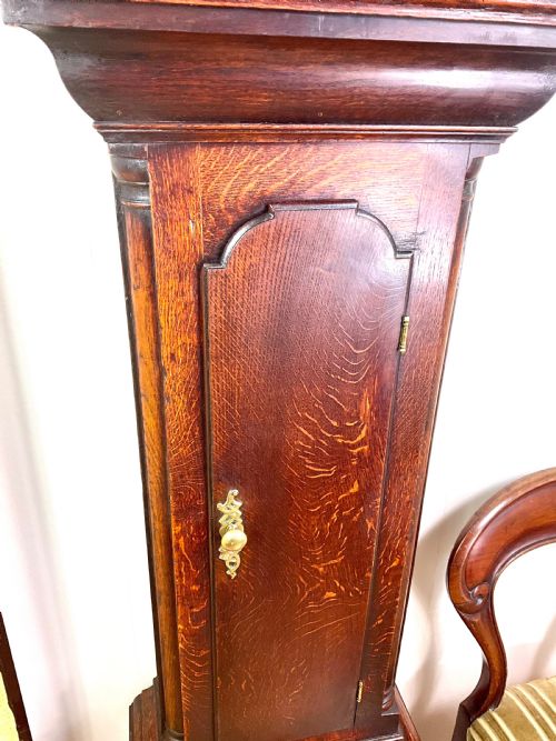 fig 2. An Early Georgian longcase clock constructed from dark oak featuring a square hood, brass dial with Roman numerals, and an arched trunk door.