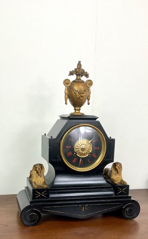 fig 1. A black slate Egyptian Revival mantel clock featuring a central dial with red Roman numerals, flanked by two gilt bronze sphinxes and topped with a gilt urn finial.