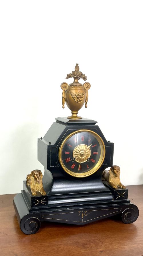 fig 2. A black slate Egyptian Revival mantel clock featuring a central dial with red Roman numerals, flanked by two gilt bronze sphinxes and topped with a gilt urn finial.