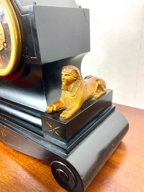 fig 3. A black slate Egyptian Revival mantel clock featuring a central dial with red Roman numerals, flanked by two gilt bronze sphinxes and topped with a gilt urn finial.