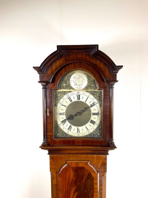 fig 1. An antique mahogany longcase clock featuring a flame mahogany trunk door, arched hood, and brass dial with Roman numerals.