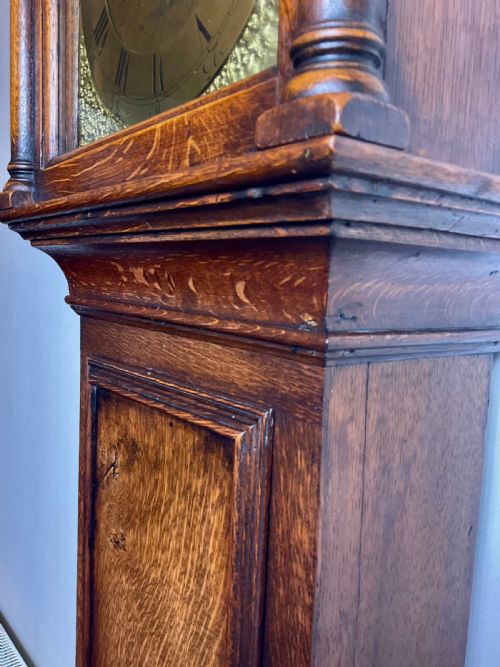 fig 1. An Early Georgian oak longcase clock featuring a square brass dial with spandrels, flanked by turned columns, and a tall wooden trunk set on bracket feet.