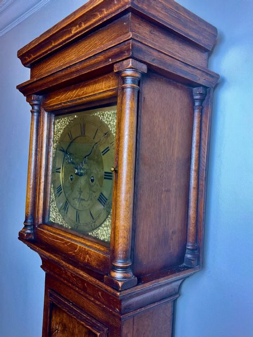 fig 2. An Early Georgian oak longcase clock featuring a square brass dial with spandrels, flanked by turned columns, and a tall wooden trunk set on bracket feet.