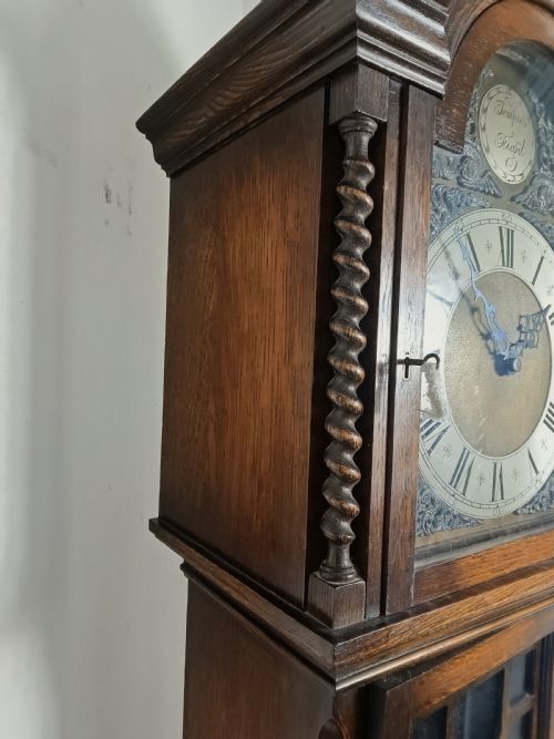 fig 1. A tall dark oak longcase clock featuring an arched bonnet, barley twist side columns, a brass dial with Roman numerals, and a glazed trunk door displaying three brass weights and pendulum.