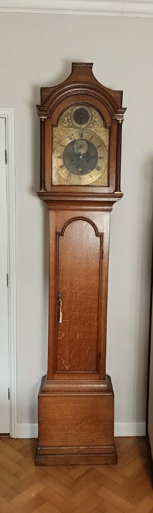 fig 1. An Early Georgian floor-standing oak longcase clock featuring a carved arch hood, brass dial with strike and silent selector, and a slim trunk door.